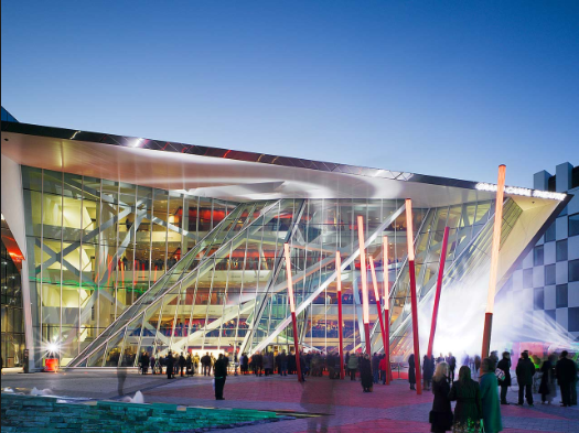 Modern architectural building with crowd gathering outside at dusk, featuring striking geometric glass design.