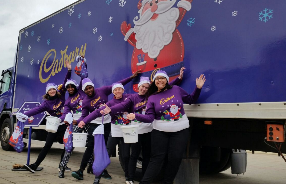 Group in festive Cadbury sweaters poses happily in front of a decorated truck with Santa.