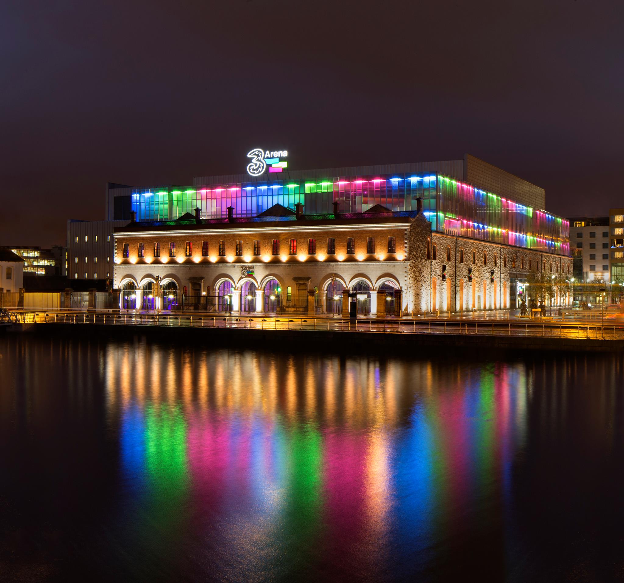 Vibrant night view of a lit-up arena with colorful reflections on water, enhancing the cityscape's ambiance.