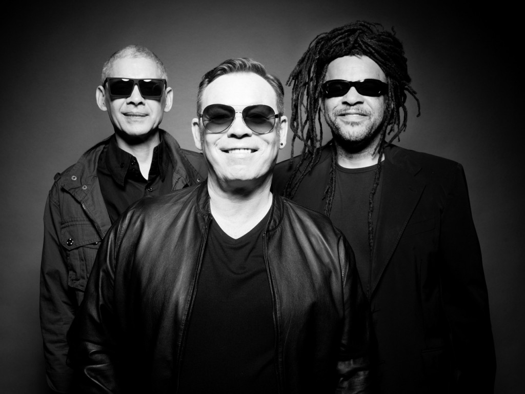 Three men in sunglasses smiling, dressed in black, posing against a dark background in a monochrome image.