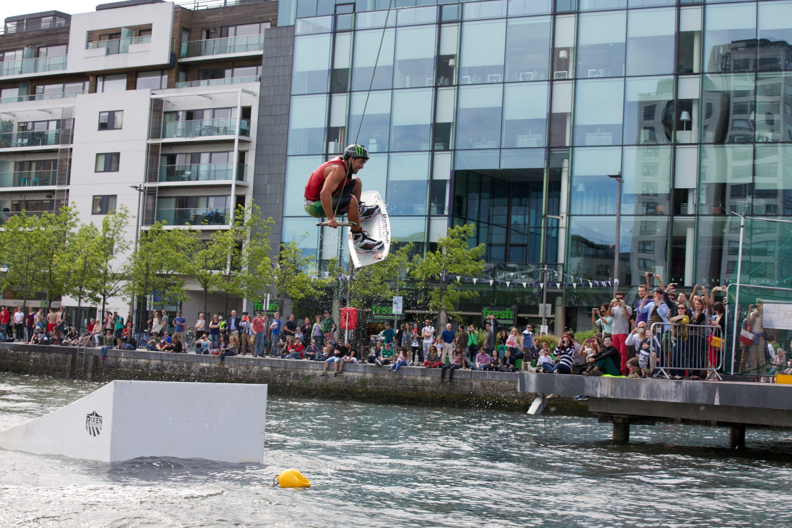 Wakeboarder performs aerial trick over water in urban setting with crowd watching on a sunny day.