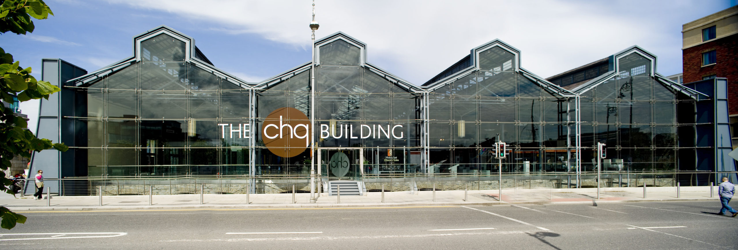 Modern glass-fronted building with The CHQ Building sign and street view, surrounded by pedestrians and greenery.