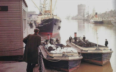 Historic harbor scene with cargo ships and workers unloading sacks on a dock.