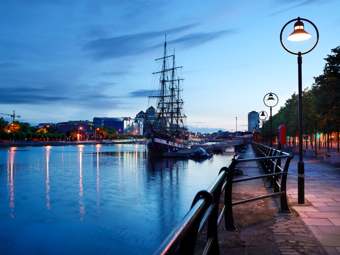 Historic sailing ship docked at a serene waterfront during evening twilight, with city lights illuminating the riverbank.
