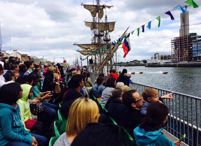 Crowd watching a tall ship event by the riverside, colorful flags flying, urban skyline in background.