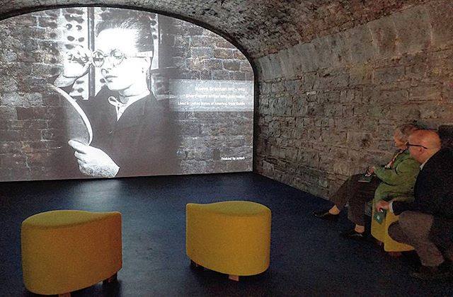 Visitors watching a black and white film in a stone-walled room with modern seating.