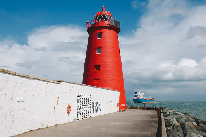 Bright red lighthouse on a pier with a ship in the ocean and a cloudy blue sky background.