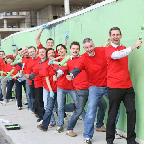 Group of volunteers in red shirts painting a wall green, smiling and holding paint rollers in an outdoor setting.