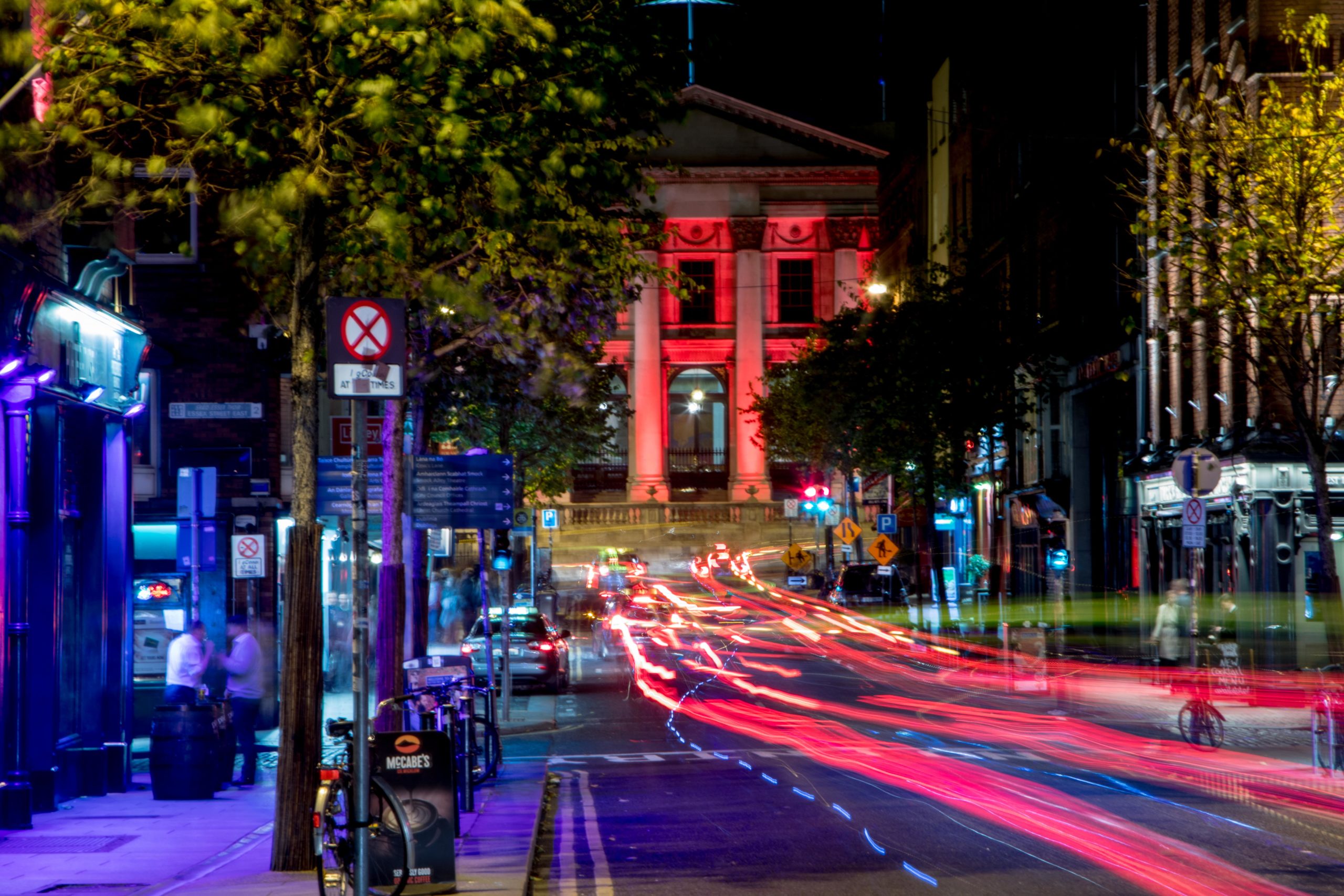 Vibrant city street at night with colorful light trails and historic building illuminated in red.