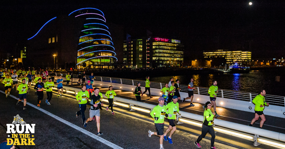 Night marathon runners cross a lit city bridge, illuminated buildings in the background.
