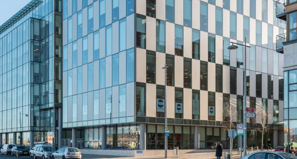 Modern office building with glass facade in urban setting, reflecting blue sky.