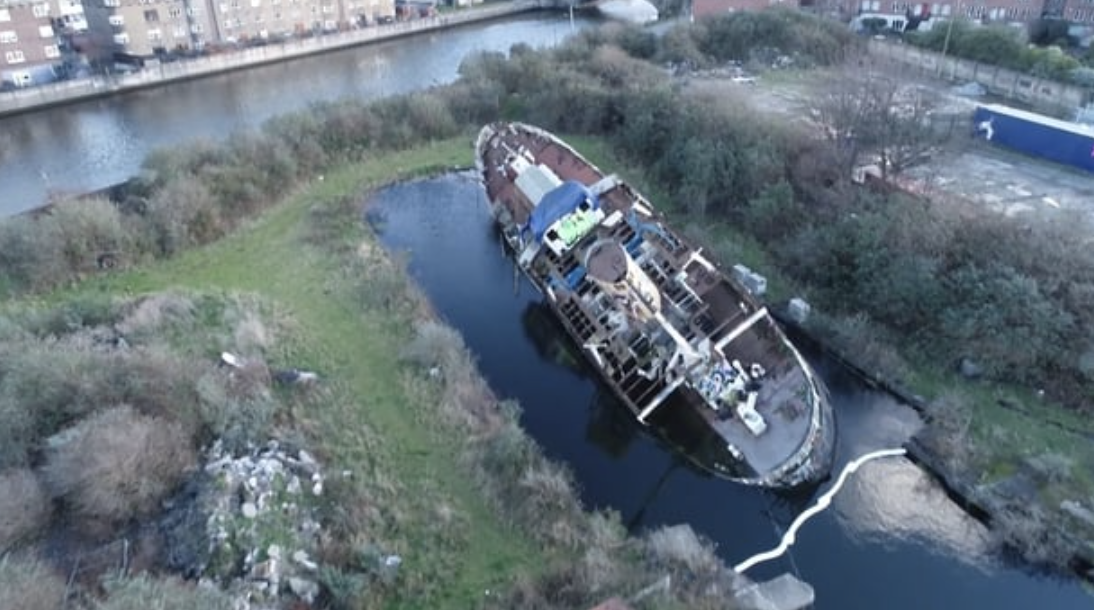 Aerial view of an abandoned ship in a small waterway surrounded by greenery and urban area.