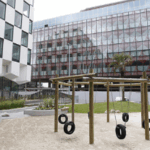 Modern office buildings with a playground featuring tire swings in the foreground.