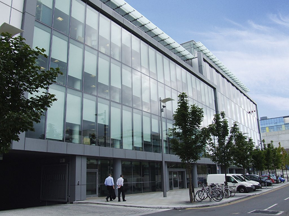 Modern office building with glass façade, people walking on sidewalk, parked cars and bicycles, trees lining the street.