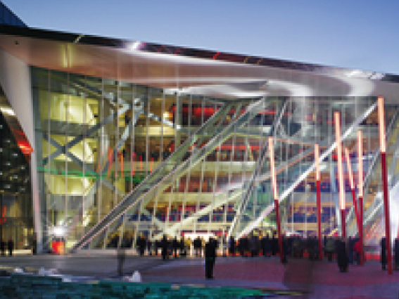 Modern glass building with illuminated facade and crowd outside at dusk.