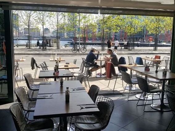 Outdoor restaurant seating overlooking a waterfront, with people dining on a sunny day.