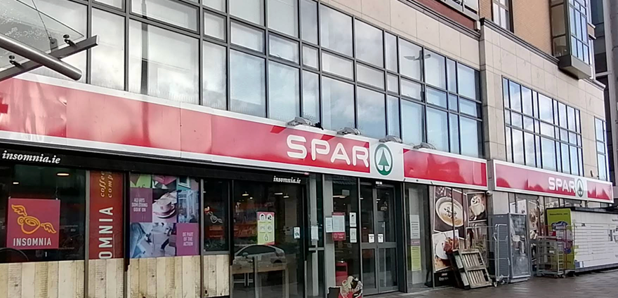 Exterior view of a Spar convenience store with glass windows and a red signboard in an urban setting.