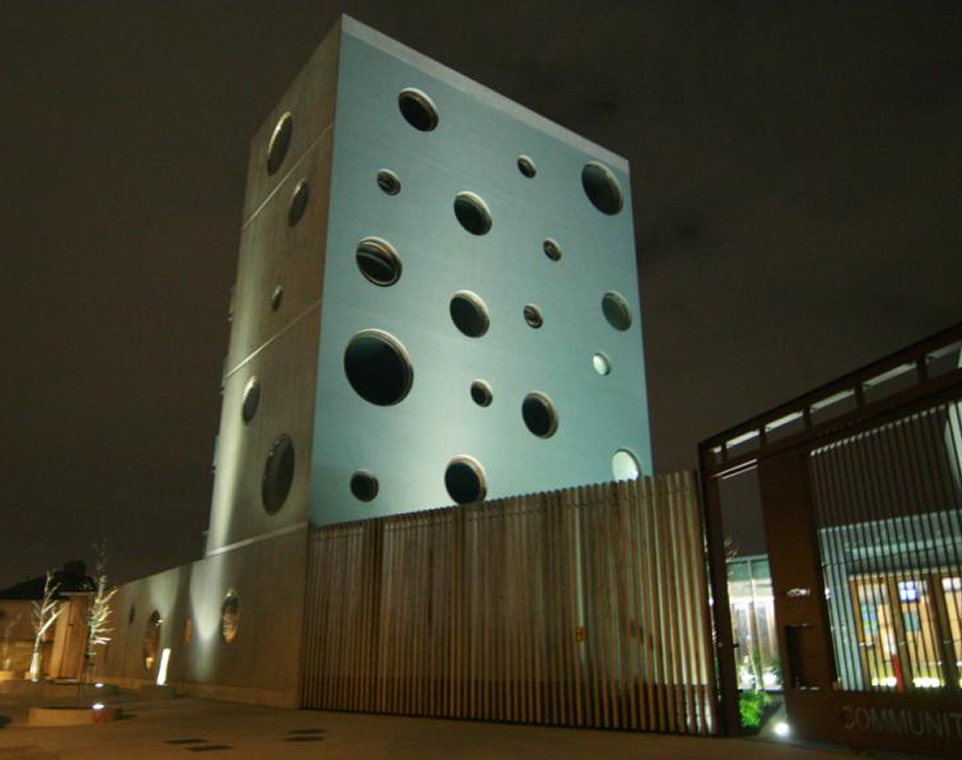 Modern building with circular windows, nighttime view. Unique architecture with illuminated facade and wooden fence.