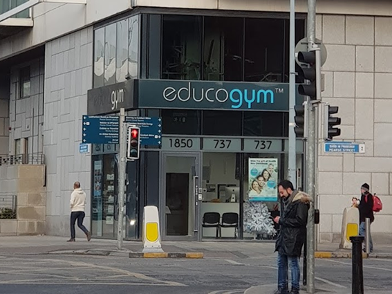 Gym entrance on a city street corner with people walking nearby, showcasing modern fitness facility.
