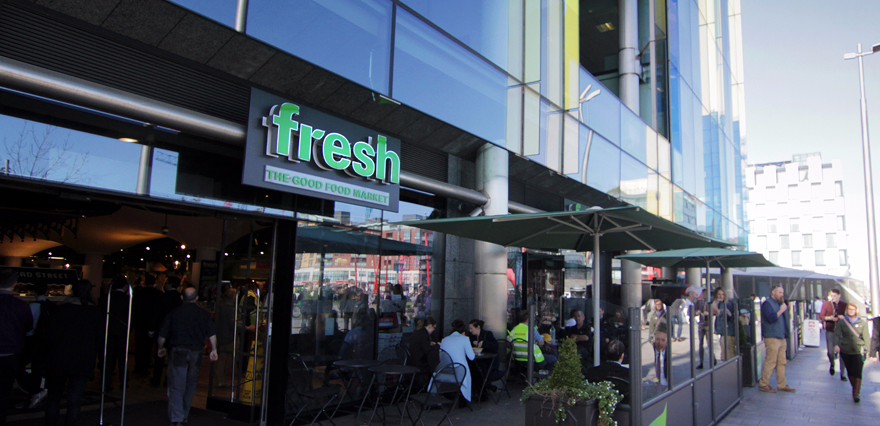 Fresh food market entrance with outdoor seating and people on a busy street.