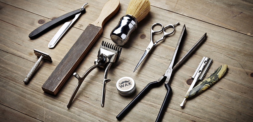 Barber tools set on wooden table, including scissors, brushes, straight razor, clippers, and shaving accessories.