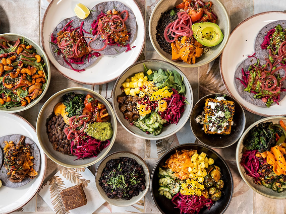 Colorful vegan dishes with tacos, bowls of beans, veggies, avocado, and garnishes, arranged on a table.