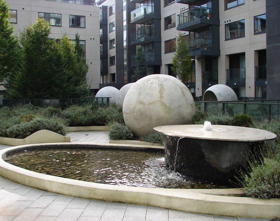 Modern courtyard with water feature, large spheres, greenery, and apartment buildings in urban setting.