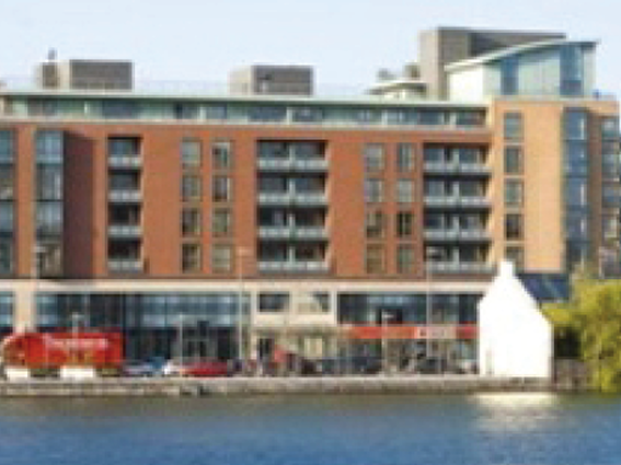 Riverside modern building with glass balconies and ground-floor shops on a sunny day.