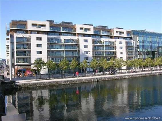 Modern waterfront apartments by a tree-lined river, reflecting in the water under a clear blue sky.