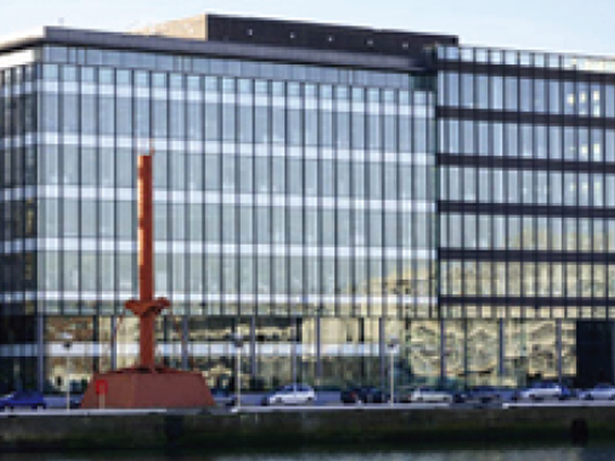 Modern glass office building with red sculpture in front, cars parked along waterfront, reflecting sunlight.