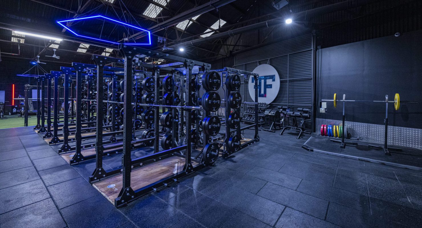 Modern gym with weightlifting equipment and neon lighting, featuring racks and colorful plates in a spacious interior.