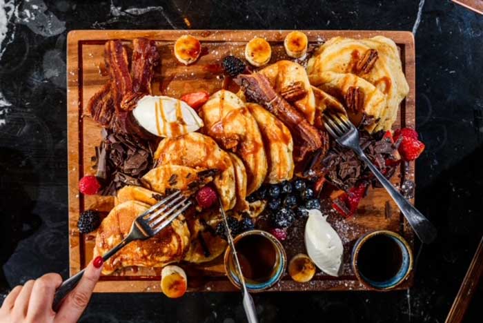 Delicious breakfast platter with pancakes, bacon, fresh berries, cream, and syrup on a wooden board.
