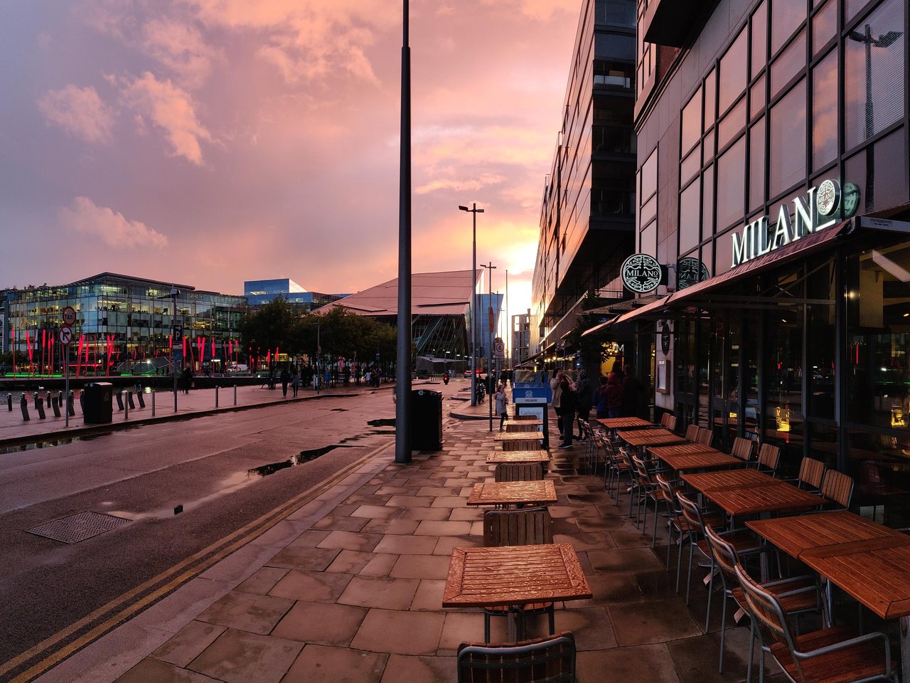 Urban street scene at sunset with restaurant patio seating, reflecting lights on wet pavement.