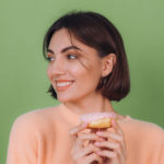 Woman smiling with a pink donut against a green background, wearing a peach sweater and hoop earrings.